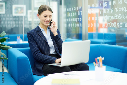 Wallpaper Mural Portrait of smiling young businesswoman using laptop in office and speaking by phone during internship, copy space Torontodigital.ca