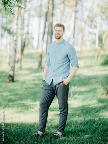 Stylish bearded man with blond hair dressed in a blue shirt posing for a photo in the park.