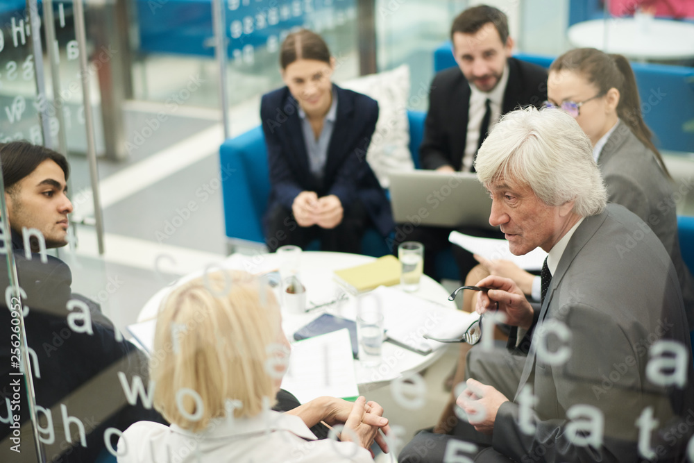 Fototapeta premium Group of business people discussing project sitting in circle during meeting, shot from behind glass, copy space