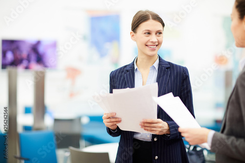 Waist up portrait of smiling young businesswoman talking to colleague while standing in office at work, copy space