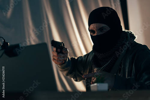 Aggressive criminal in mask pointing with finger and aiming gun at laptop screen photo