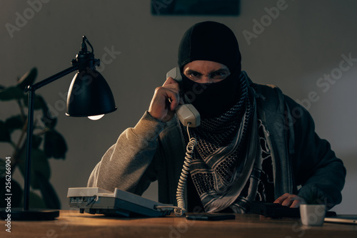 Angry terrorist in mask sitting at table with lamp and talking on telephone