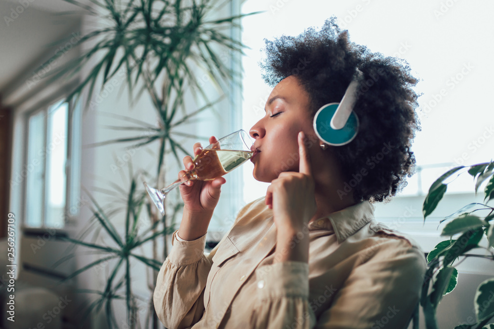 © Mediteraneo - Young beautiful african american woman relaxing and listening to music using headphone, drinking wine.
