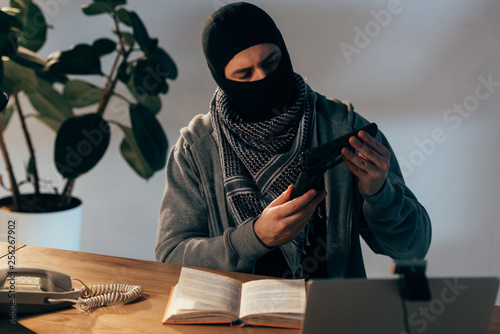 Pensive terrorist in black mask looking at gun in room photo