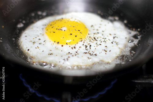 Egg sunny side up with salt and pepper, frying in a pan on the stove.