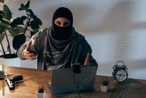 Angry terrorist in black mask gesturing at webcam in room