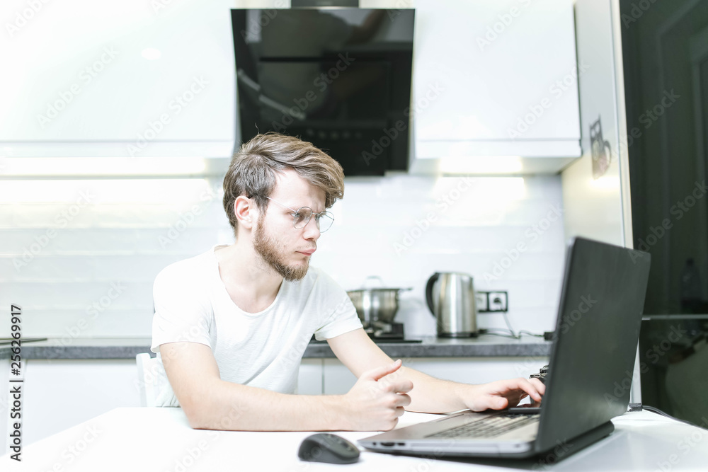 bearded young man wearing glasses in the kitchen using a laptop, searching the Internet for furniture and home appliances bearded young man wearing glasses in the kitchen using a laptop, searching the Internet for furniture and home appliances