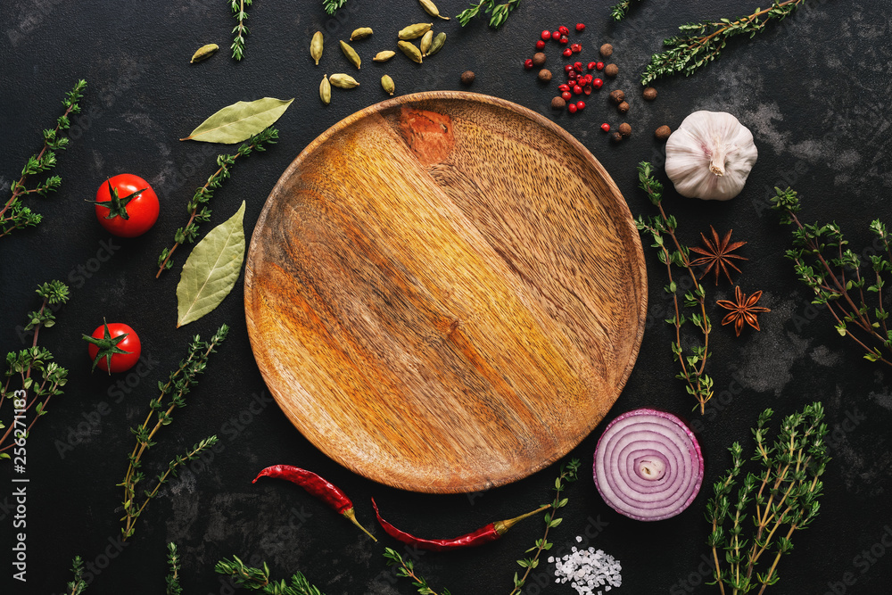 Empty wooden plate and frame of spices, herbs and vegetables on a dark stone background. Top view, flat lay.