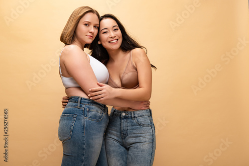 pensive overweight young woman and smiling slim asian girl hugging while looking at camera isolated on beige, body positivity concept