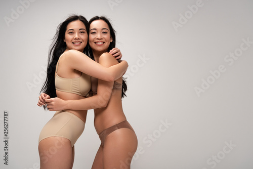 two pretty multicultural young women hugging while posing at camera isolated on grey, body positivity concept
