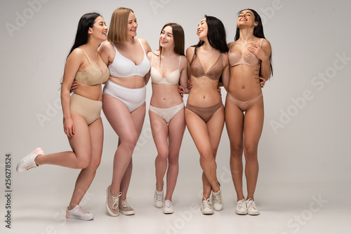 five cheerful multicultural young women in lingerie hugging while posing at camera, body positivity concept