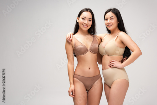 two attractive multiethnic women in lingerie hugging and looking at camera isolated on grey, body positivity concept