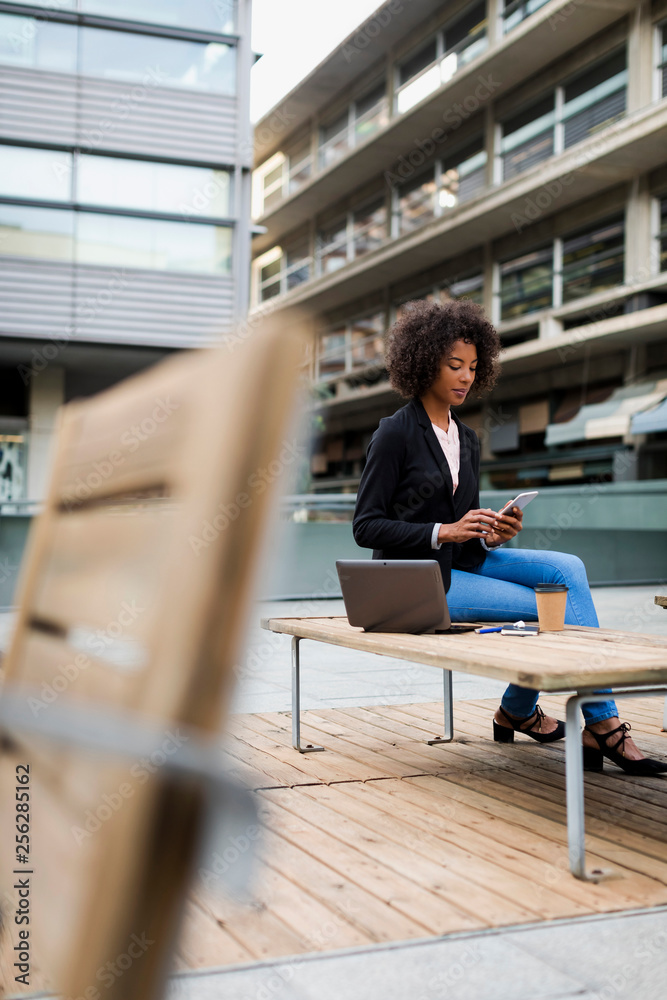 Businesswoman with laptop and coffee to go sitting on terrace looking at smartphone Businesswoman with laptop and coffee to go sitting on terrace looking at smartphone