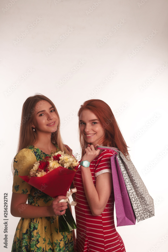 The portrait of two young smiling lades redhead and brunette with a bouquet and shoping bags in there hands isolated over white background posing The portrait of two young smiling lades redhead and brunette with a bouquet and shoping bags in there hands isolated over white background posing