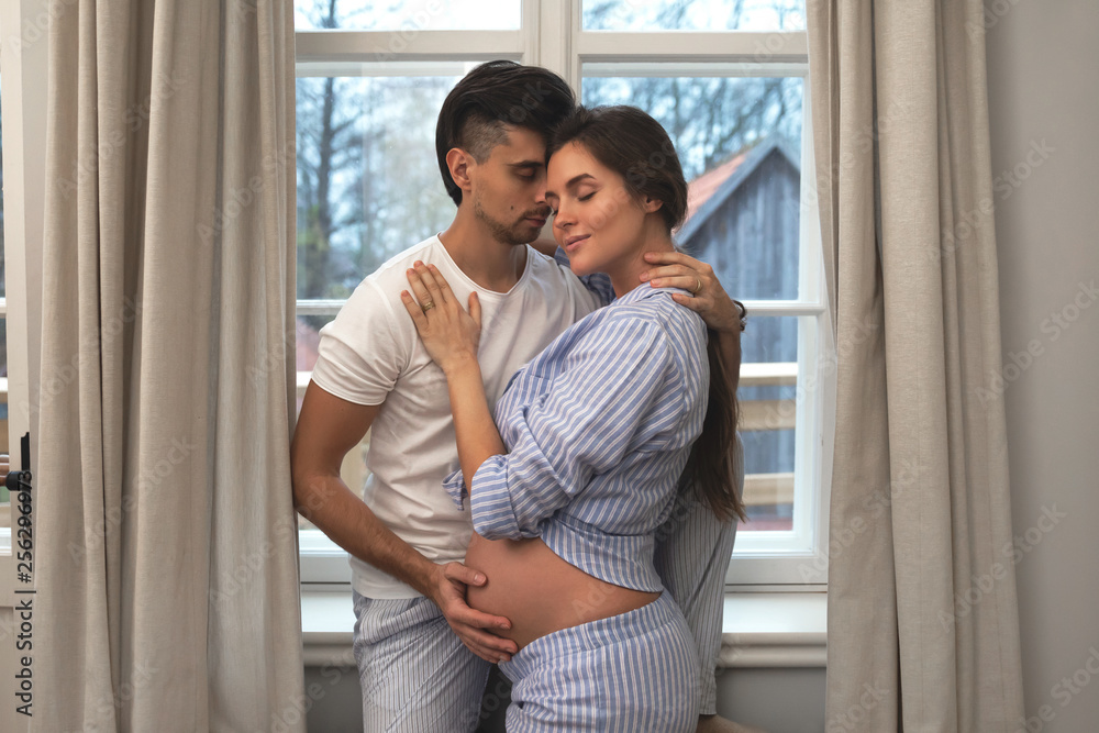 Husband and his pregnant wife in embrace beside the window Husband and his pregnant wife in embrace beside the window