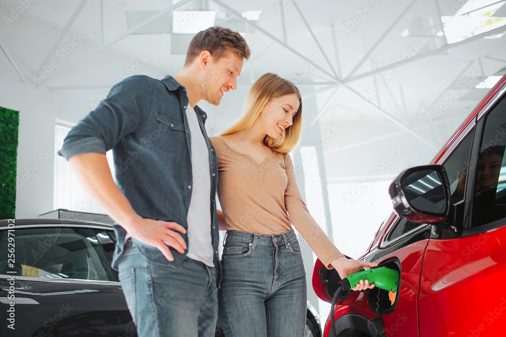 Young smiling couple buying first electric car in the showroom. Woman charging modern eco-friendly vehicle with the power cable supply plugged in