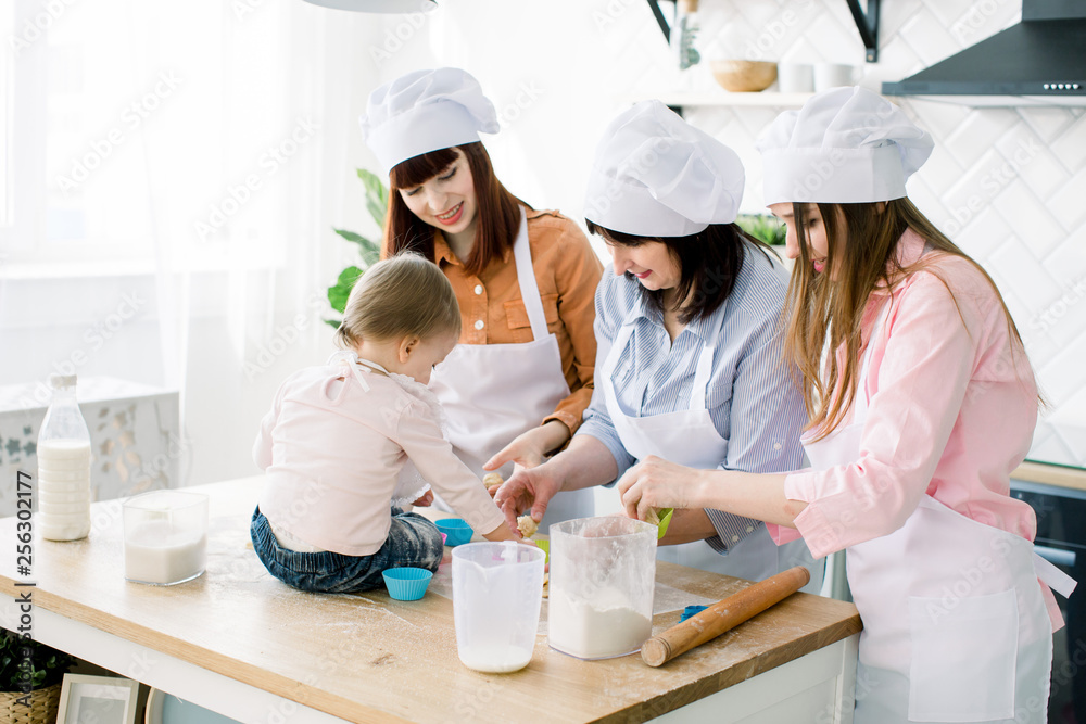 Cute little girl sitting on the table and her beautiful mother, aunt and grandmother in aprons are preparing dough for muffins and smiling while baking in kitchen at home. Mothers Day concept Cute little girl sitting on the table and her beautiful mother, aunt and grandmother in aprons are preparing dough for muffins and smiling while baking in kitchen at home. Mothers Day concept
