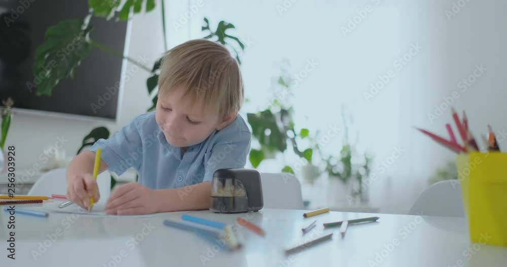 Smiling boy in blue shirt draws on paper with a pencil while sitting at the table in the living room Smiling boy in blue shirt draws on paper with a pencil while sitting at the table in the living room