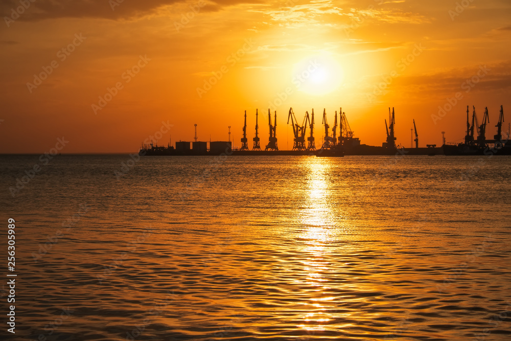 Beautiful landscape with fiery sunset sky and sea. Harbor on the coast during sunrise. Cranes silhouettes against fiery, orange and red sky.