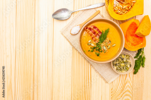 Homemade autumn hot pumpkin cream soup with smocked bacon and seeds. Raw pumpkin, fresh chives, dill, salt in spoon, vintage linen cloth on wooden background, top view copy space.