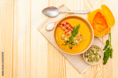 Homemade autumn hot pumpkin cream soup with smocked bacon and seeds. Raw pumpkin, fresh chives, dill, salt in spoon, vintage linen cloth on wooden background, top view.