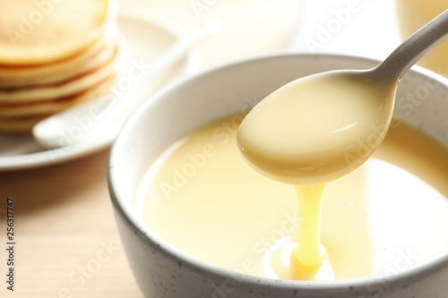 Spoon of pouring condensed milk over bowl on table  closeup with space for text. Dairy products