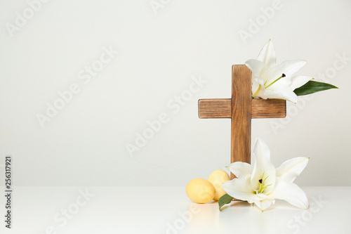 Wooden cross, Easter eggs and blossom lilies on table against light background, space for text
