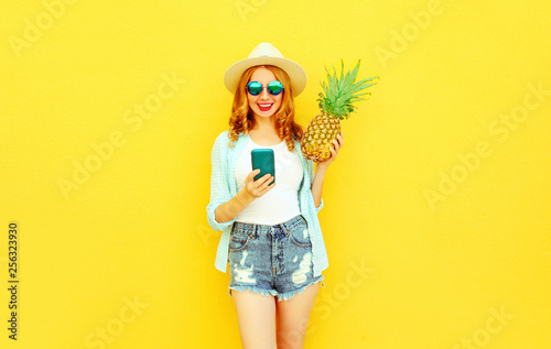 Happy smiling woman holding smartphone, pineapple in summer straw hat, shorts on colorful yellow wall background