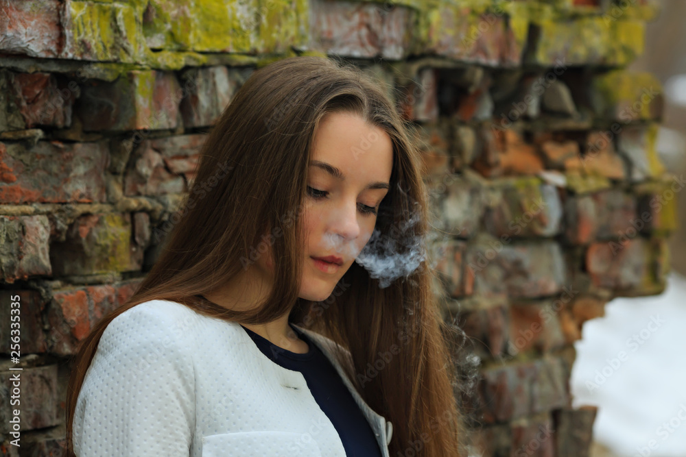Naklejka premium Vape teenager. Young pretty white girl in white cardigan smoking an electronic cigarette opposite destroyed brick wall on the street in the spring. Bad habit.