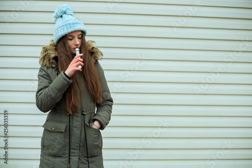 Vape teenager. Young pretty white girl in blue cap and dark green jacket smoking an electronic cigarette opposite modern brown background on the street in the winter. Bad habit.