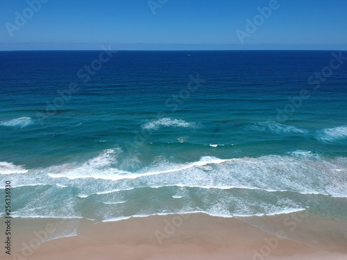 Beautiful blue pacific ocean off of an Australian beach with gentle waves, a surfer and a lone sailboat on the horizon.