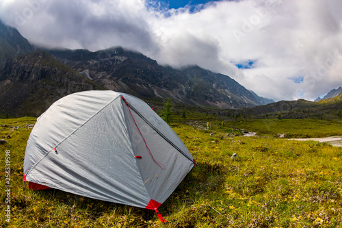 Gray tent stands in a meadow in the mountains near the lake in a thunderstorm
