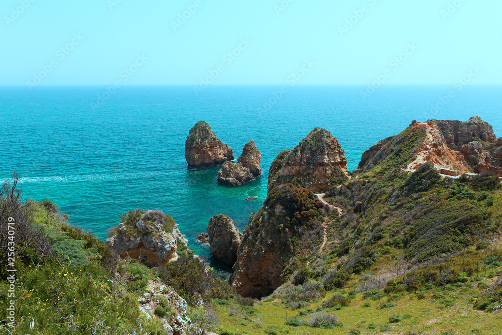 Pedestrian trails between cliffs with scenic views on turquoise sea waters and coastal line. Tourists explore grottoes on motorboat in Algarve, Portugal. Pedestrian trails between cliffs with scenic views on turquoise sea waters and coastal line. Tourists explore grottoes on motorboat in Algarve, Portugal.