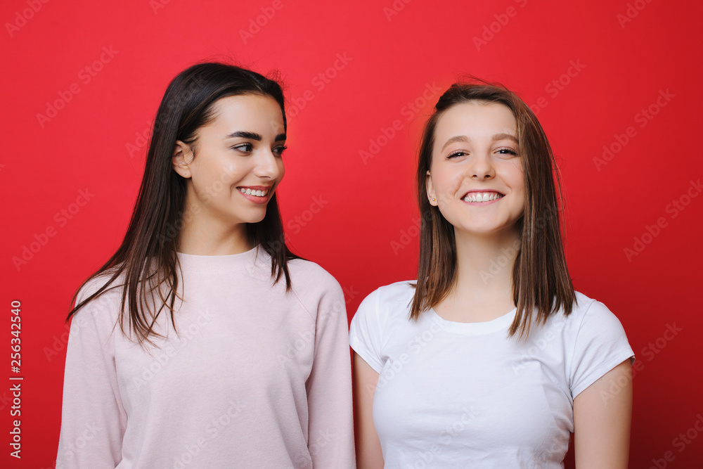 Lovely young caucasian woman looking at camera laughing while another woman is looking at her laughing in front of a red studio wall. Lovely young caucasian woman looking at camera laughing while another woman is looking at her laughing in front of a red studio wall.
