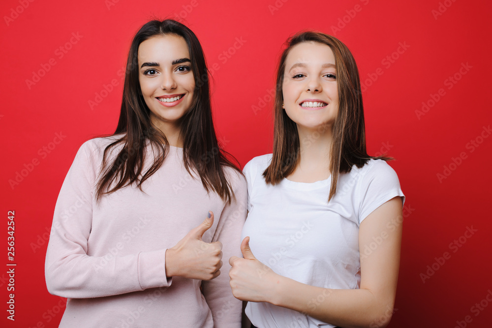 Two beautiful young attractive girlfriends showing that all is good while looking at camera laughing against red background. Two beautiful young attractive girlfriends showing that all is good while looking at camera laughing against red background.