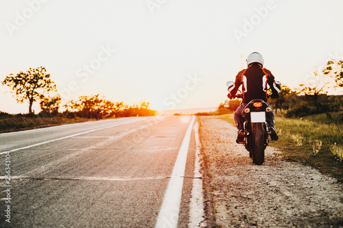 Back view of a equiped biker sitting on his motorcycle stopping on the edge of the road while traveling by motorcycle.