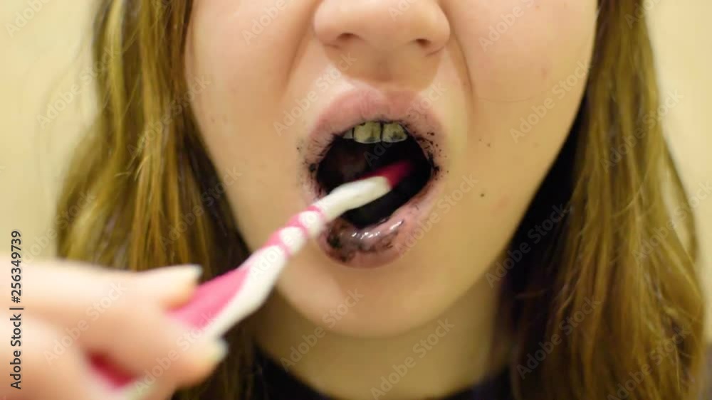 A young girl brushes her teeth with black paste. Paste with charcoal for teeth whitening. The reflection in the mirror. A young girl brushes her teeth with black paste. Paste with charcoal for teeth whitening. The reflection in the mirror.