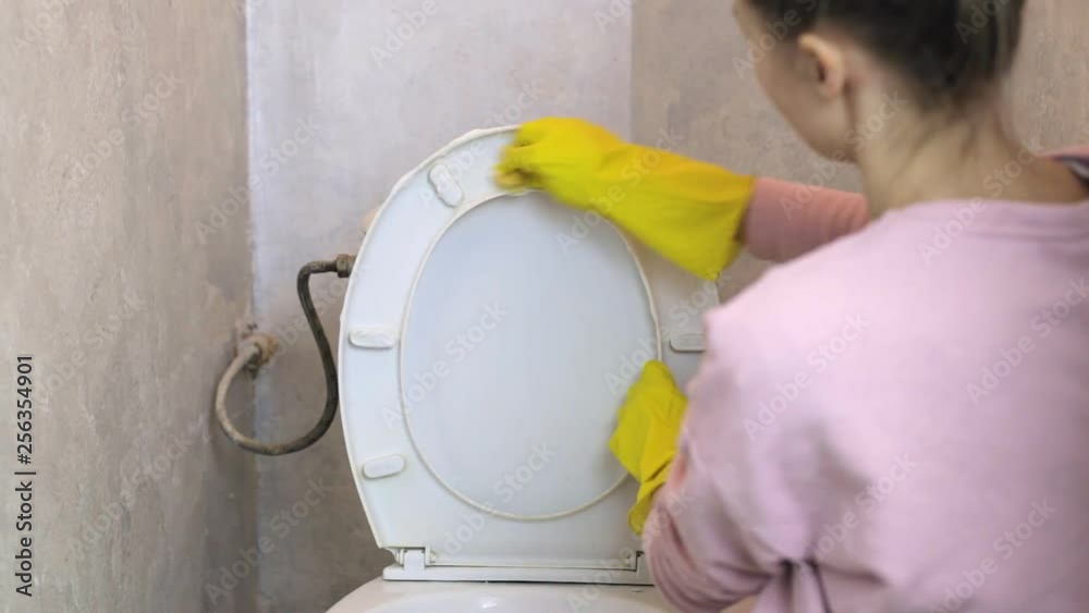 Woman cleans white toilet bowl with a sponge in yellow rubber gloves in home clothes Woman cleans white toilet bowl with a sponge in yellow rubber gloves in home clothes