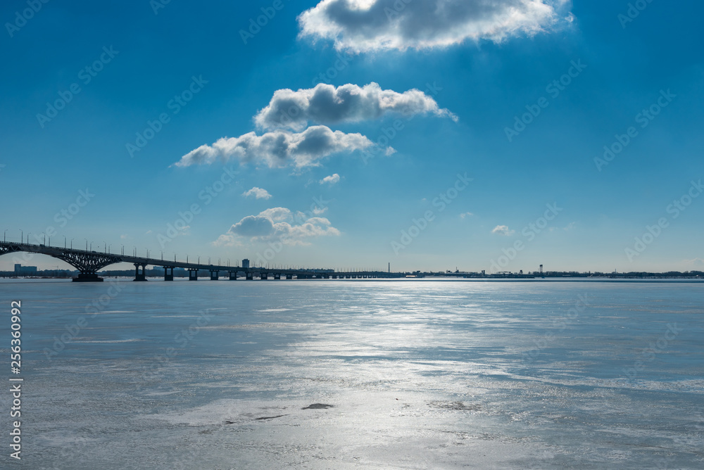 Fototapeta premium Beautiful winter landscape with a bridge over a frozen river and clouds in the blue sky