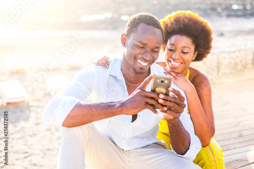 Cheerful black race diversity beautiful young couple use technology device modern phone outdoor with sun light in background - smile and happiness for cute man and woman together friends