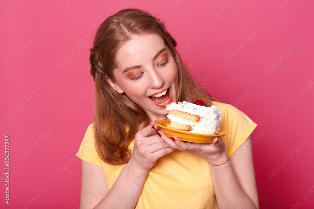 Photo of young female in casual yellow t shirt, ready for eating huge piece of cake, looks at tasty pastry, isolated on rose background, studio portrait. Culinary, people and lifestyle concept. Photo of young female in casual yellow t shirt, ready for eating huge piece of cake, looks at tasty pastry, isolated on rose background, studio portrait. Culinary, people and lifestyle concept.