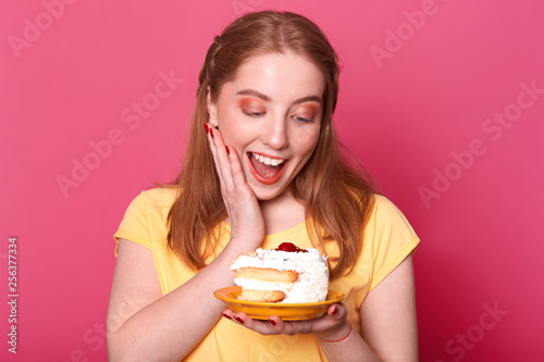 Close up portrait of satisfied pleased girl with light brown hair, holds huge piece of tasty cake, keeps her mouth open, full of enjoyment, dressed in casual yellow t shirt, poses in photo studio. photo