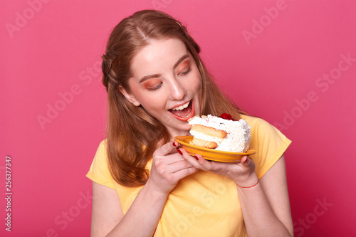 Photo of young female in casual yellow t shirt  ready for eating huge piece of cake  looks at tasty pastry  isolated on rose background  studio portrait. Culinary  people and lifestyle concept.