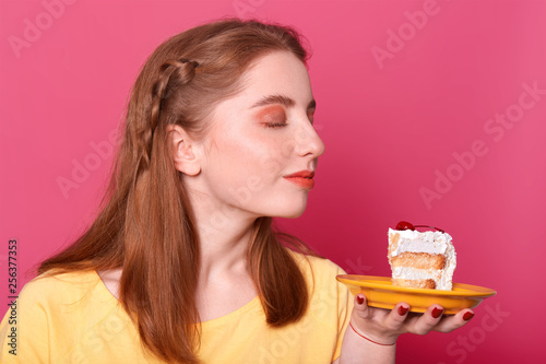 Horizontal shot of good looking young female  model sniffs tasty dessert  concentared at huge piece of cake  wants to eat delicious pastry  poses in photo studio isolated over pink background.