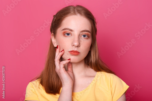 Indoor portrait of attractive good looking girl with long straight hair, has serious facial expression, keeps hand on cheek, has bright red manicure, wears casual yellow t shirt. People concept.