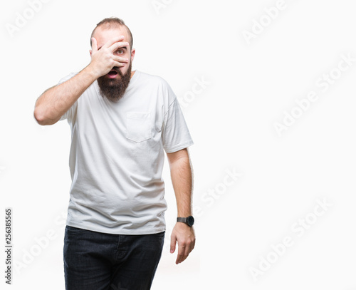 Young caucasian hipster man wearing casual t-shirt over isolated background peeking in shock covering face and eyes with hand, looking through fingers with embarrassed expression.