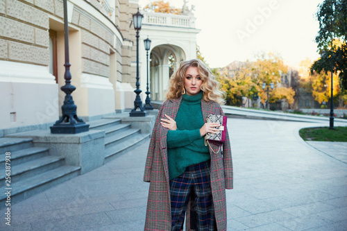 woman in trendy outfit walking in street.Outdoor waist up portrait of young beautiful woman with curly hair, looking aside,wearing warm long coat,green sweater and high heels.Odessa Opera theatre.