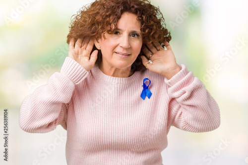 Middle ager senior woman wearing changeable blue color ribbon awareness over isolated background Trying to hear both hands on ear gesture, curious for gossip. Hearing problem, deaf © Krakenimages.com