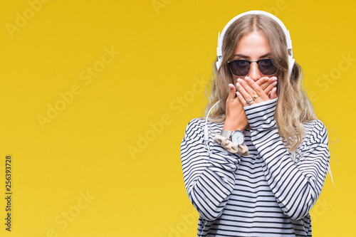 Beautiful young blonde woman wearing headphones and sunglasses over isolated background shocked covering mouth with hands for mistake. Secret concept.
