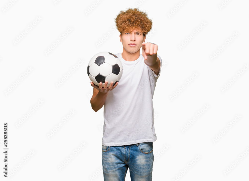 Young handsome man holding soccer football ball pointing with finger to the camera and to you, hand sign, positive and confident gesture from the front